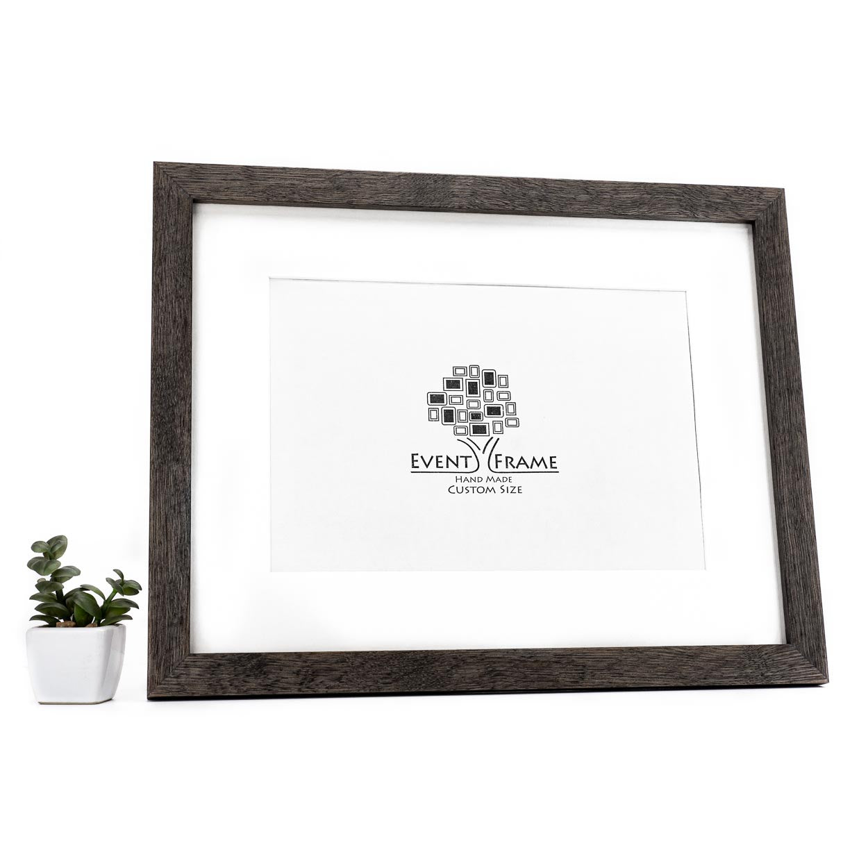 A black solid oak hardwood photo frame with a white matte border, displaying an artwork logo, placed on a white surface with a small potted plant to the side.