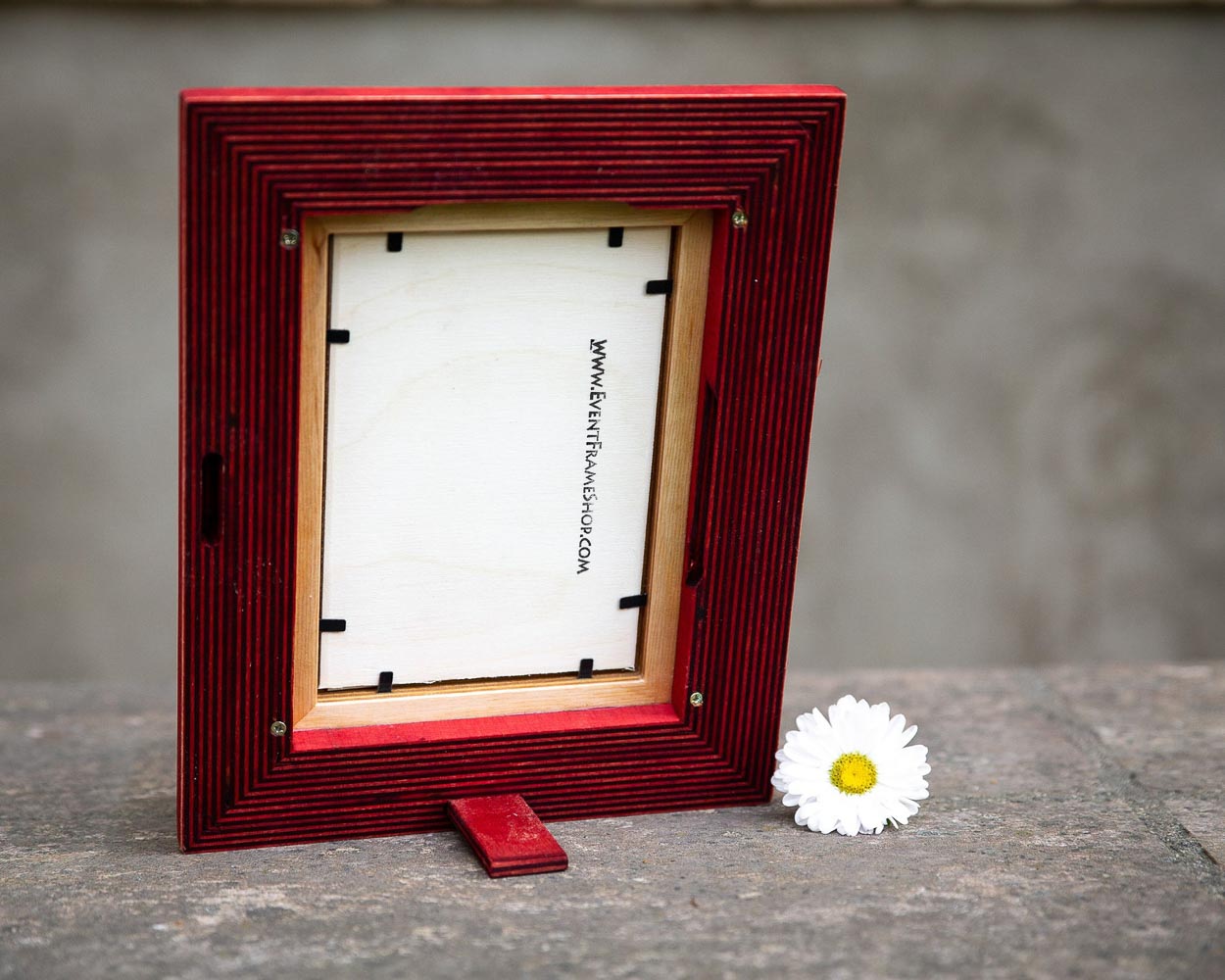 A rustic design picture frame with a red exterior and natural wood interior, displayed with a white artwork piece and a small flower beside it.